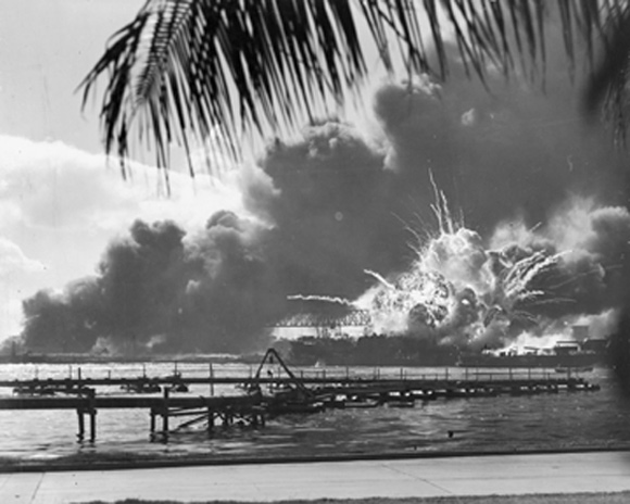 Photographic image of Pearl Harbor in smoke. The image is taken at some distance and is framed by a palm tree and undamaged pavement.